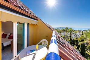 A balcony with a glass door leading to a bedroom, a table with a glass of orange juice, and a view of the sea and palm trees. - Bahia de Marbella | Bright Beachside Apartment with Pool in Marbella (Marbella)