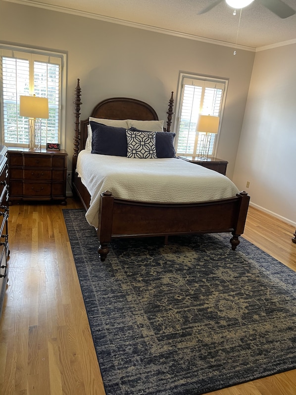 Une chambre à coucher avec un lit en bois, deux tables de chevet, une commode, un tapis à motifs et une fenêtre avec des stores.