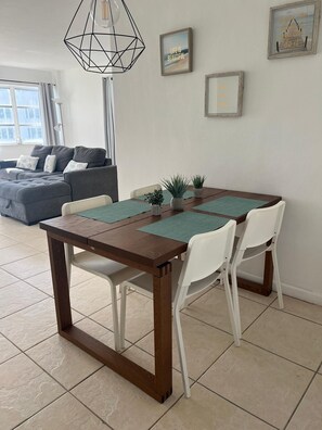 Uma área de refeições com mesa de madeira e cadeiras brancas, luminária pendente geométrica e um sofá ao fundo.