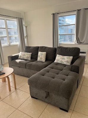 Um sofá modular cinzento com almofadas estampadas, uma mesinha redonda e uma janela com cortinas.
