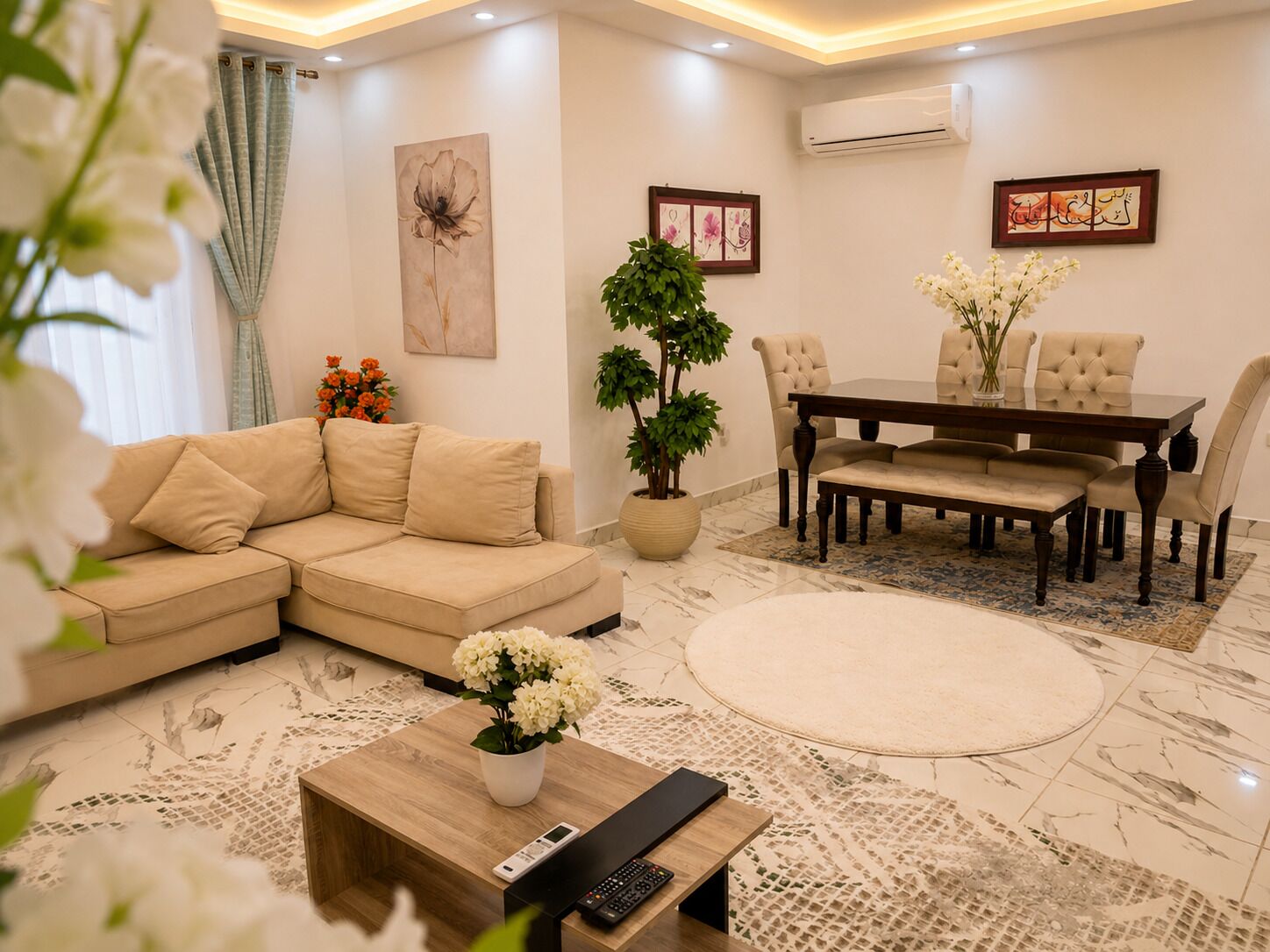 Una sala moderna con un sofá beige, una mesa de comedor con sillas, una mesa de centro con un control remoto y una planta en maceta.