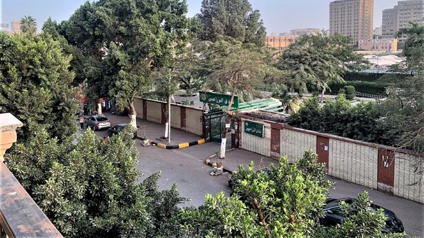 Un área de estacionamiento cerrada con un letrero verde, rodeada de árboles y edificios al fondo.