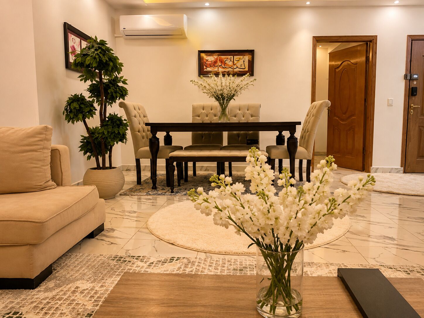 Una sala de estar con sofá, una mesa de comedor con sillas, una planta en maceta y un jarrón con flores.