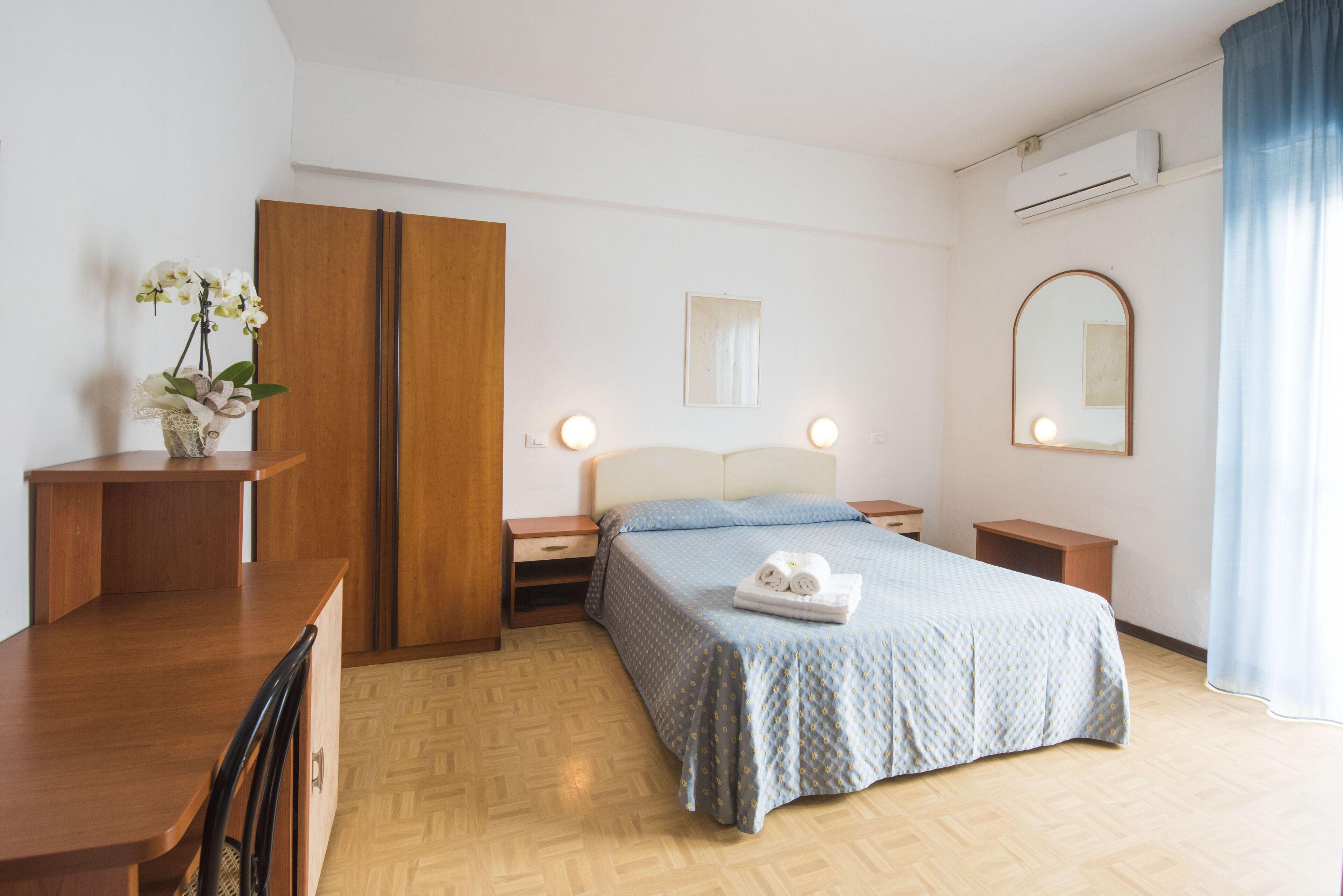 A hotel room with a bed, bedside tables, a desk, a wardrobe, a mirror, and an air conditioning unit.
