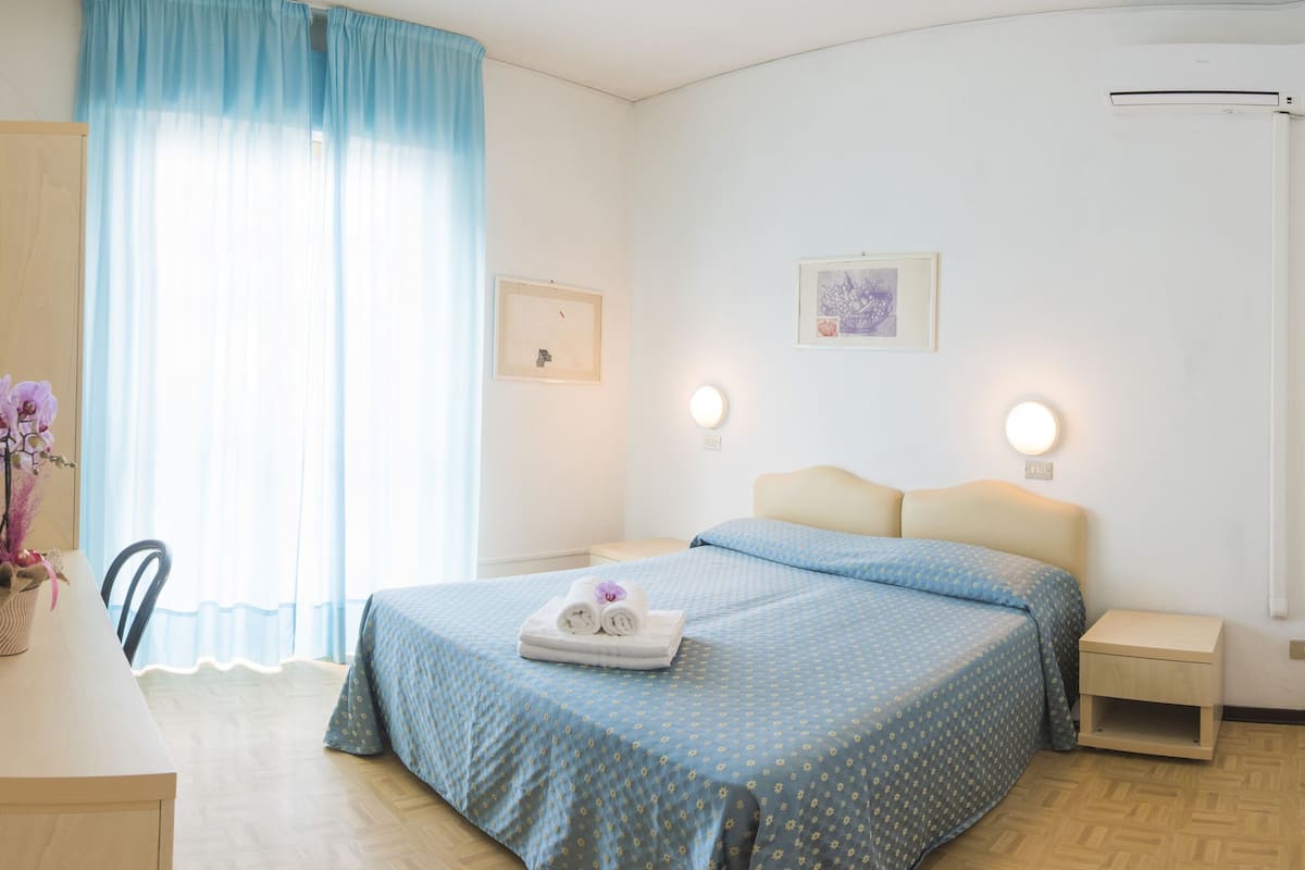 A hotel room with a bed, bedside table, nightstand, dresser, mirror, and a window with curtains.
