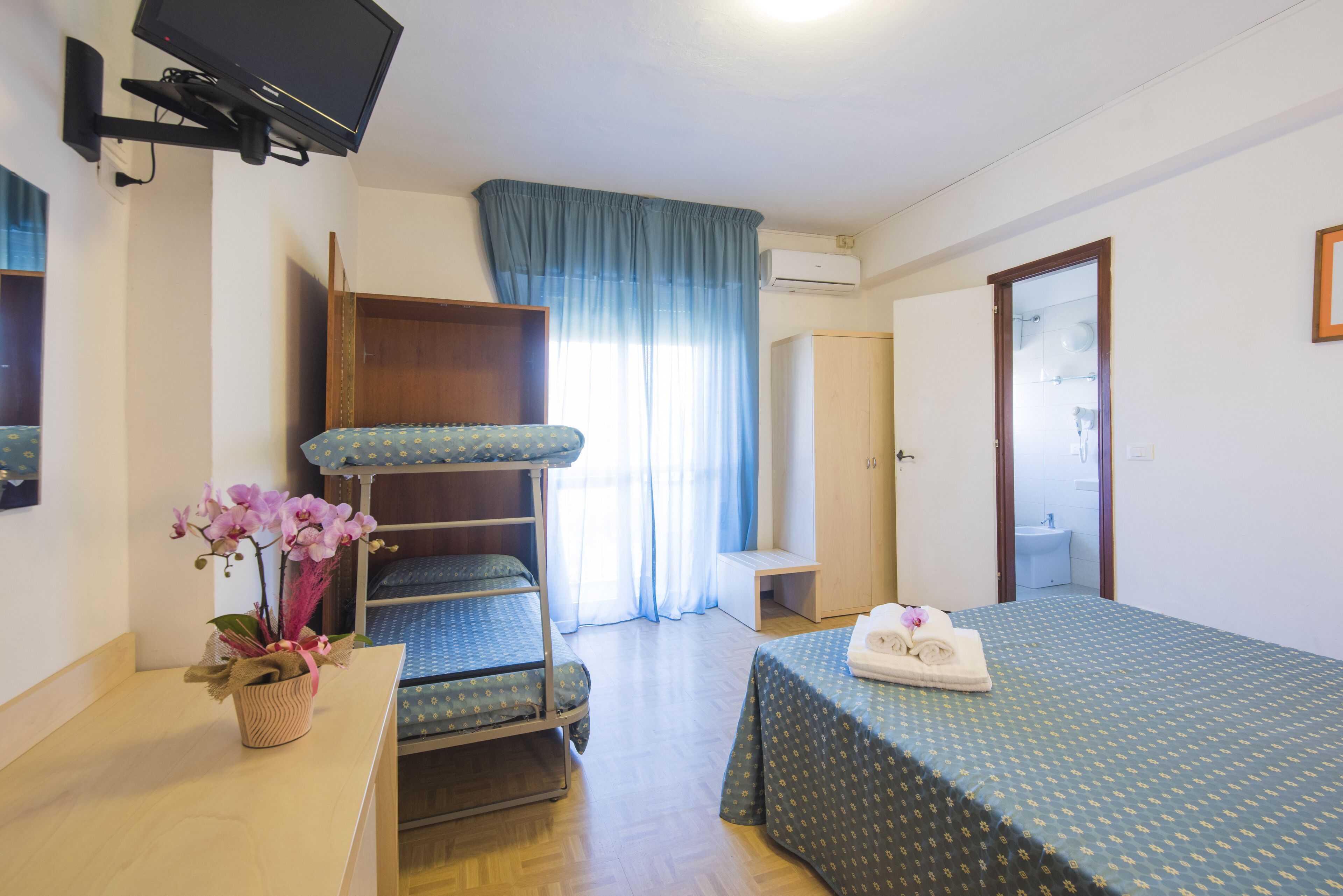 A hotel room with bunk beds, a desk with a potted orchid, a bed with a green patterned bedspread, a television mounted on the wall, and a bathroom visible through an open door.