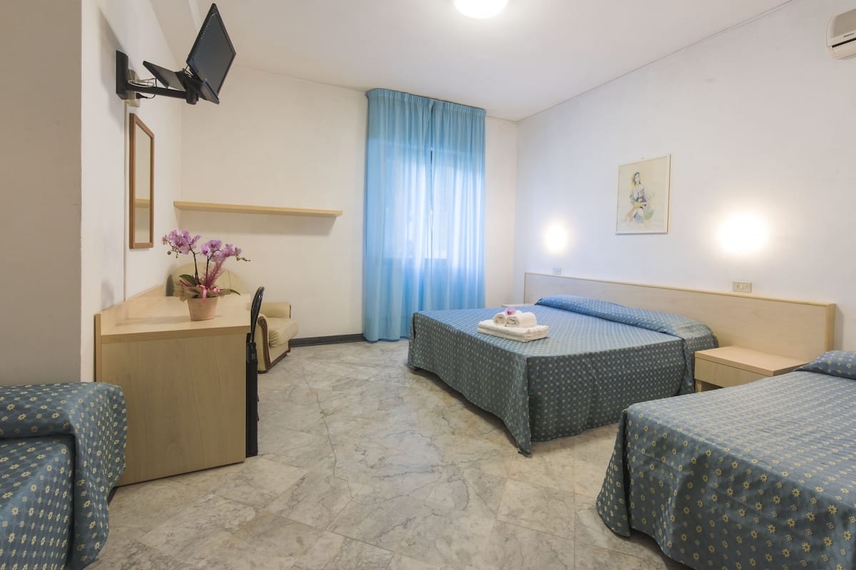 A hotel room with two single beds, a desk with a chair, a small table with a vase of flowers, a TV mounted on the wall, and a window with blue curtains.