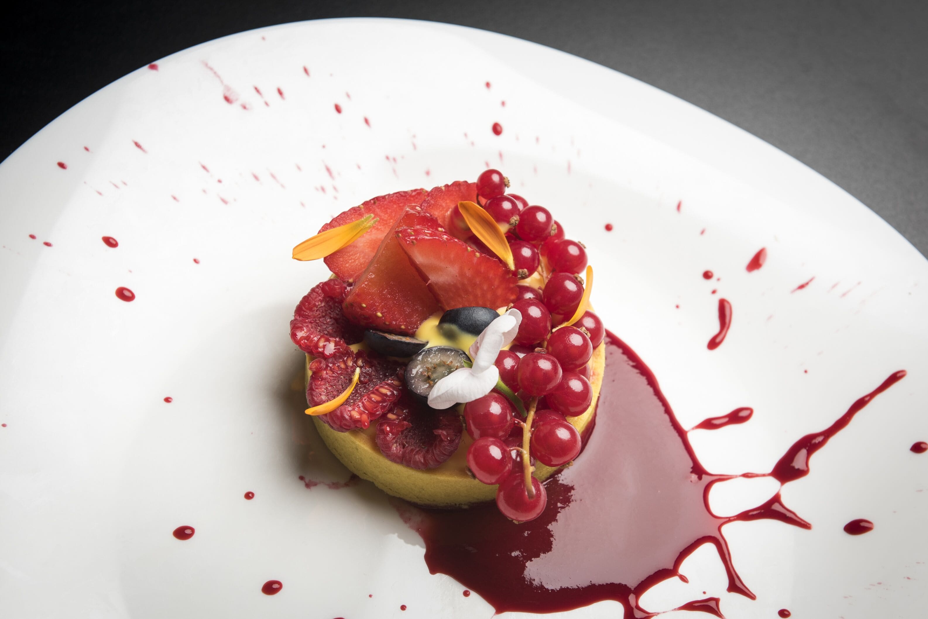 Postre en un plato blanco con fruta roja y un centro de crema blanca, rodeado de salpicaduras de salsa roja.