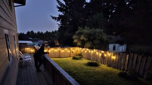 Een houten terras met een leuning, een gazon en een houten omheining met lichtjes.