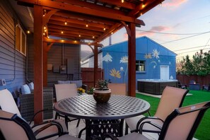 Une terrasse couverte avec une table ronde et des chaises, une plante en pot, et un bâtiment bleu orné de peintures murales florales en arrière-plan.