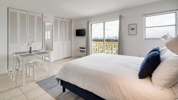 A bedroom with a bed, a desk, a chair, a TV, and a balcony with a city view. - Modern Apartment| City Views + Easy Beach Access (Miami)