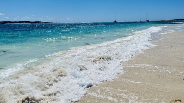 Ein Strand mit klarem türkisblauem Wasser, sanften Wellen und einem entfernten Segelboot.