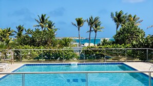 Ein rechteckiger Pool mit glasklarer, blauer Wasseroberfläche, umgeben von einem Metallzaun. Im Hintergrund sind Palmen und ein Blick auf das Meer mit Booten zu sehen.