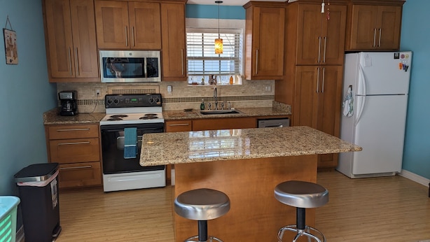 A modern kitchen with wooden cabinets, a granite countertop, a built-in oven, a microwave, a refrigerator, and two bar stools. - Lovely Guest Cottage min from Perdido Key Beaches (Pensacola)
