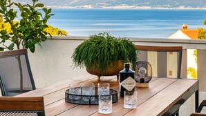 Une table en bois avec des chaises, une plante en pot et une bouteille de gin Hendricks sur un balcon donnant sur une vue côtière.