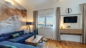 Un salon moderne avec un canapé bleu, une table basse en bois, un téléviseur fixé au mur et une grande fenêtre avec des rideaux.