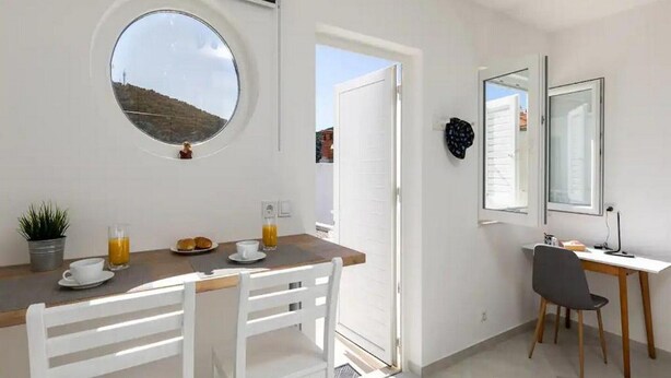 Een kleine kamer met een rond raam, een tafeltje met ontbijtartikelen, een bureau met een stoel, en een deur naar buiten.
