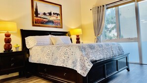 Une chambre à coucher avec un grand lit, deux lampes de chevet, une table de nuit, une tête de lit en bois, un tableau au mur et une fenêtre avec des rideaux.