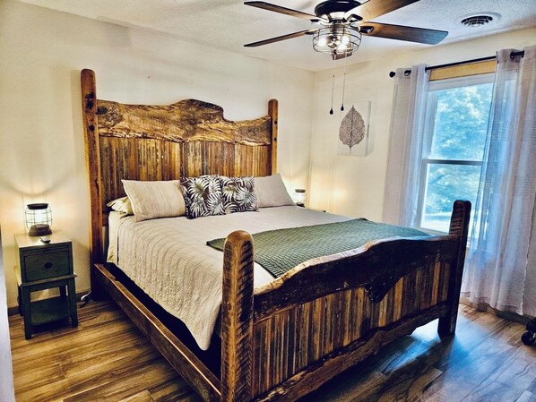 Uma cama de madeira com cabeceira, uma mesa de cabeceira com abajur e uma janela com cortinas.