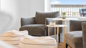 Ein modernes Wohnzimmer mit einem grauen Sofa, einem runden Couchtisch und Blick auf den Strand durch ein großes Fenster.