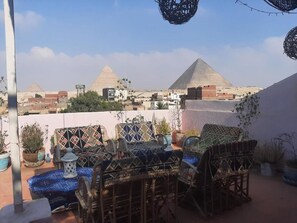 Eine Terrasse mit Korbmöbeln und Blumentöpfen, mit Blick auf Pyramiden und eine Stadtlandschaft.