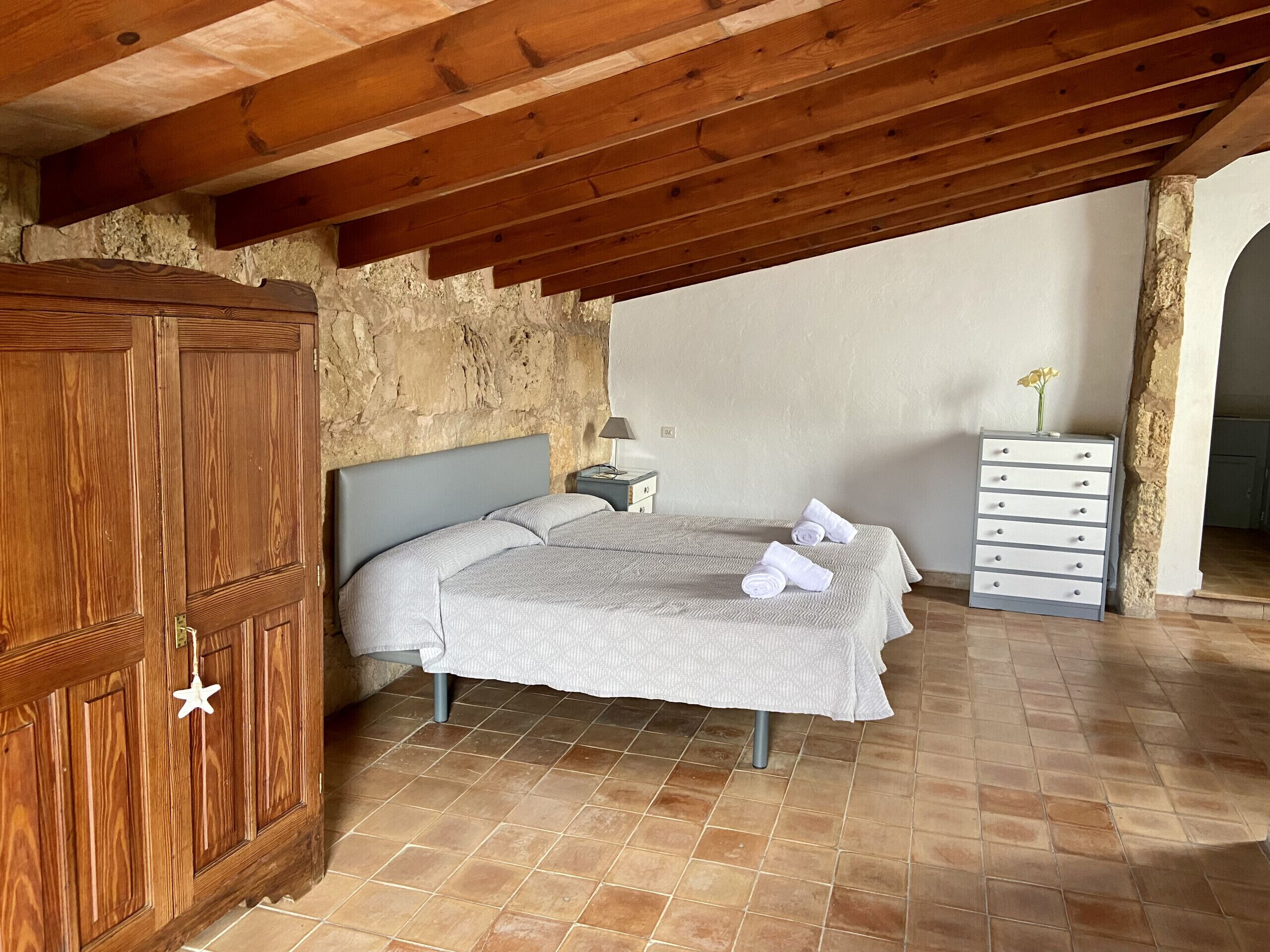 Een slaapkamer met een houten plafond, een stenen muur, een bed met een grijs hoofdeinde, een nachtkastje en een dressoir.