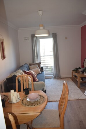 A cozy living room with a sofa, dining table, and chairs. There is a window with curtains, a hanging light fixture, and a small side table with a plant.