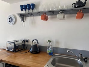 A kitchen with a toaster, kettle, sink, and a shelf with cups and glasses.