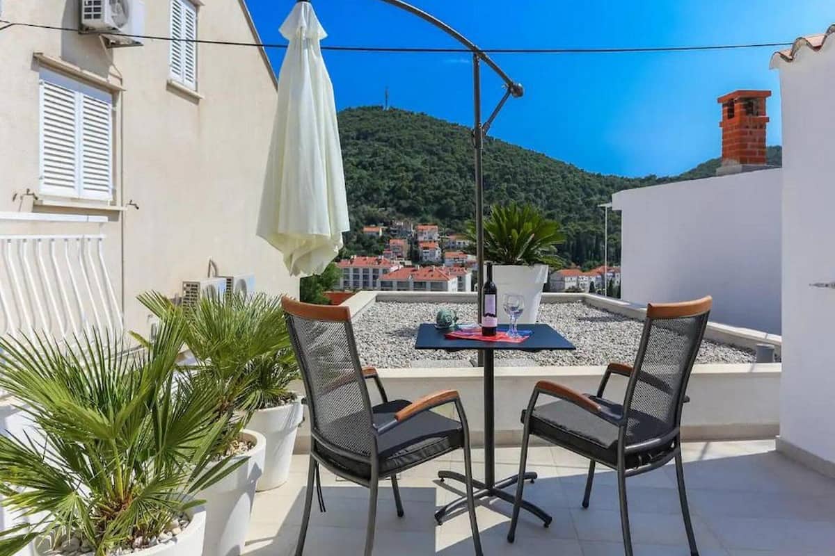 Um terraço com duas cadeiras, uma mesa, um guarda-sol branco e vista para uma vila no alto de uma colina.