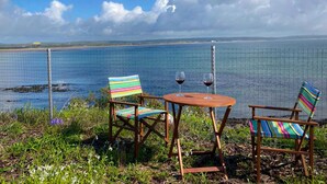 A coastal outdoor seating area with two chairs and a table, overlooking a body of water and a distant shoreline. - Ballycotton Holiday Apartment No 6 (Ballycotton)