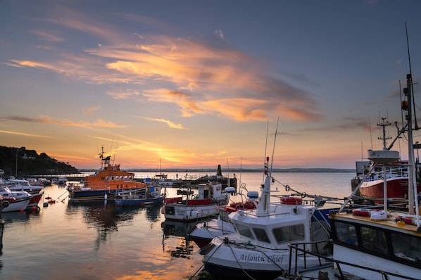 A marina with various boats docked, a lighthouse, and a sunset in the background.