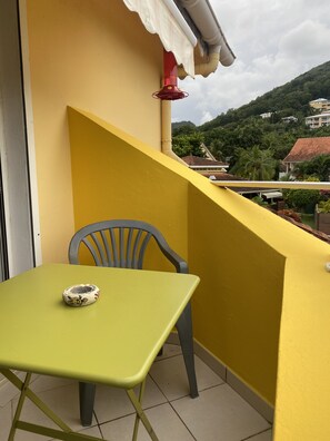 Un balcon jaune avec une table et une chaise, un tuyau rouge, et une vue sur une colline verdoyante parsemée de bâtiments.