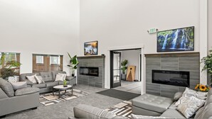 A modern living room with a grey sofa, a coffee table, and a fireplace. There are two TV screens mounted on the walls, and a large window with blinds. - Townhome 47, 3 Bed-12 Guests with indoor/Outdoor pools+Rec Center at Waterdance (Garden City)