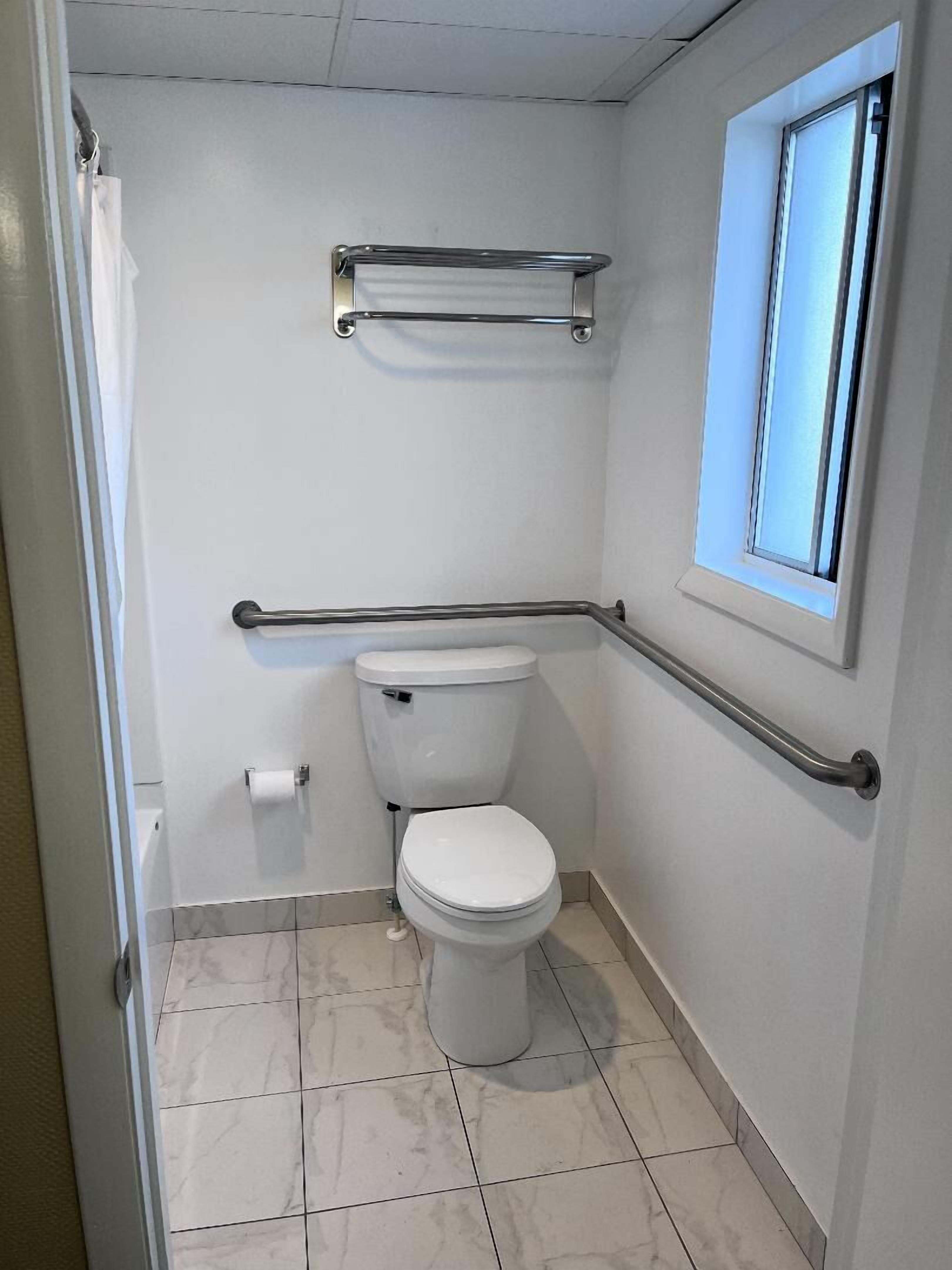 A small, clean bathroom with a toilet, grab bars, a towel rack, and a window.