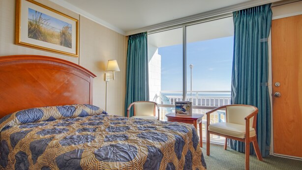 A hotel room with a large bed, a desk, two chairs, a view of the ocean, and a balcony with curtains. - Sahara Motel (Ocean City)