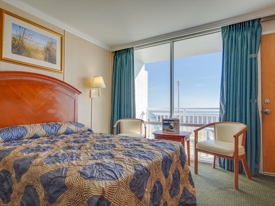Quarto de hotel com uma cama grande, uma escrivaninha, duas cadeiras, vista para o oceano e uma varanda com cortinas.