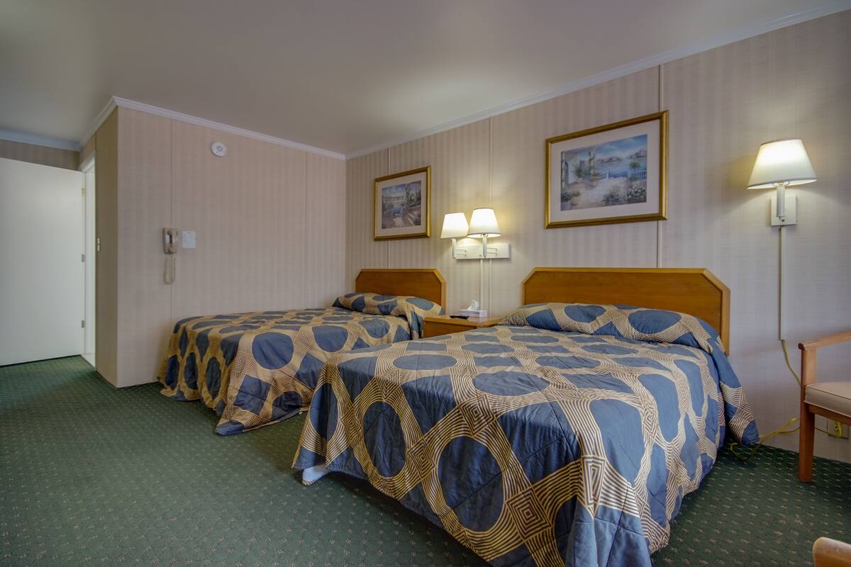 A hotel room with two beds, a desk, a chair, and a lamp.