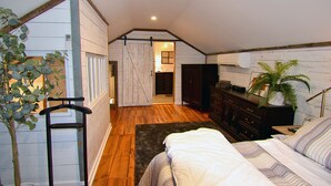A bedroom with a wooden floor, a bed with striped bedding, a dresser, a plant, and a view of a bathroom through a sliding door.