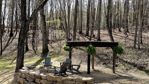 A rustic outdoor seating area with stone walls and wooden furniture, surrounded by a forest.