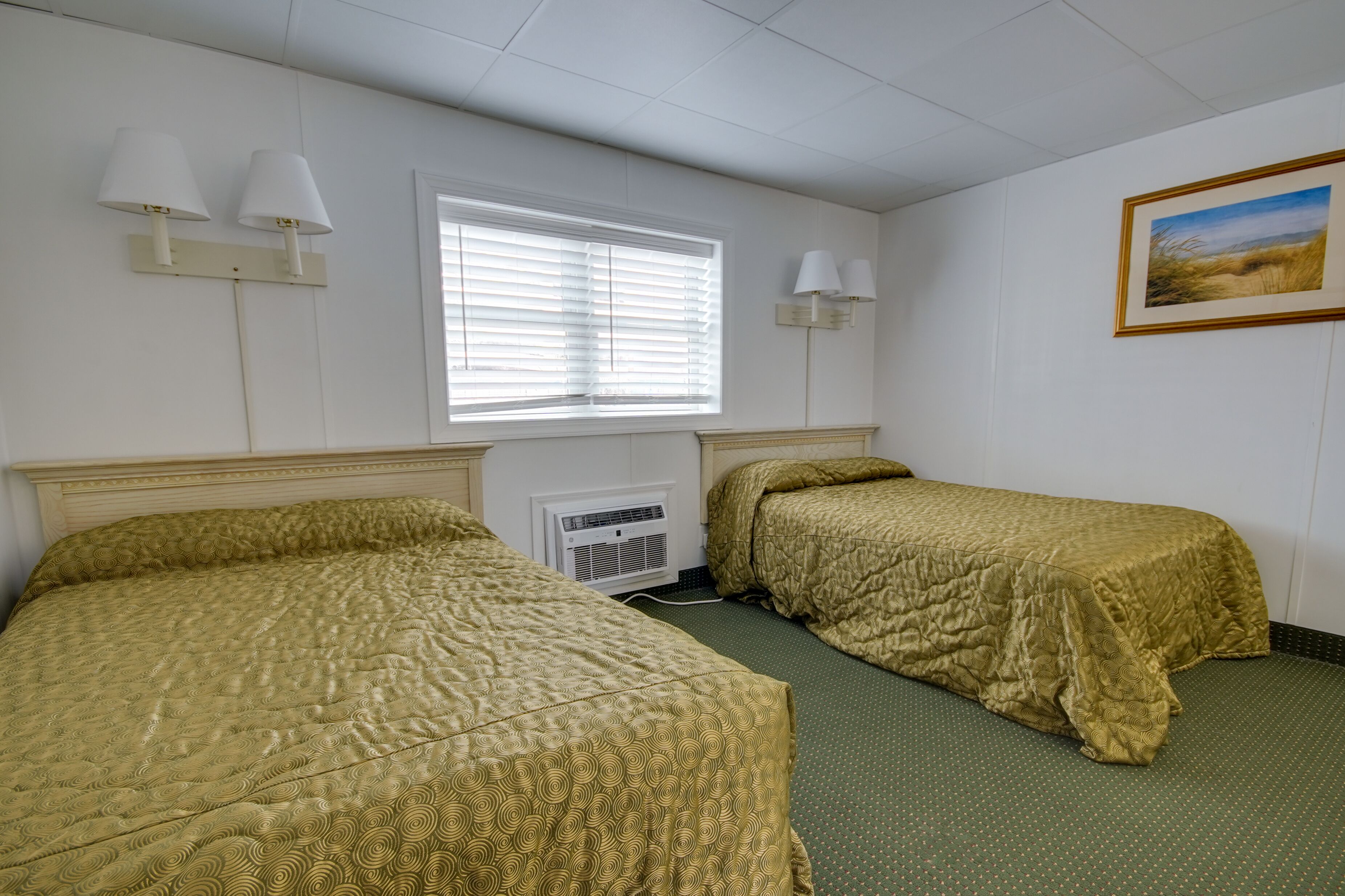 A hotel room with two single beds, a window with blinds, a wall-mounted air conditioner, and a framed picture on the wall.