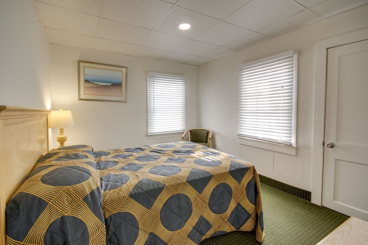 A hotel room with a bed, a chair, a painting on the wall, and two windows with blinds.