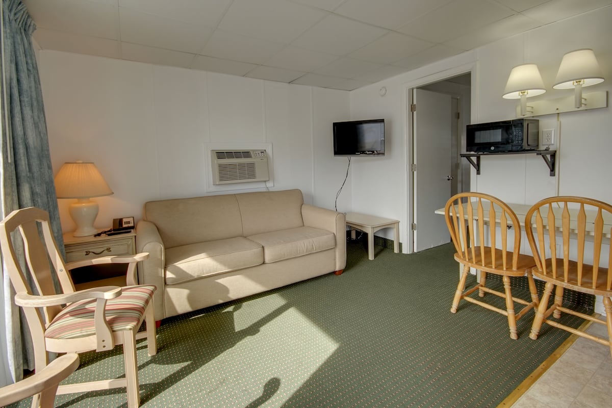 A room with a beige sofa, wooden chairs, a small table, a TV, an air conditioning unit, and a lamp.