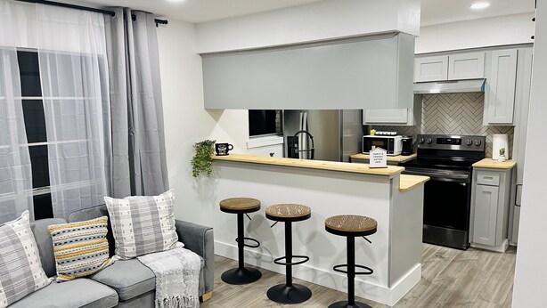 Cozinha moderna com ilha central, banquetas de bar e um sofá cinza com almofadas estampadas.