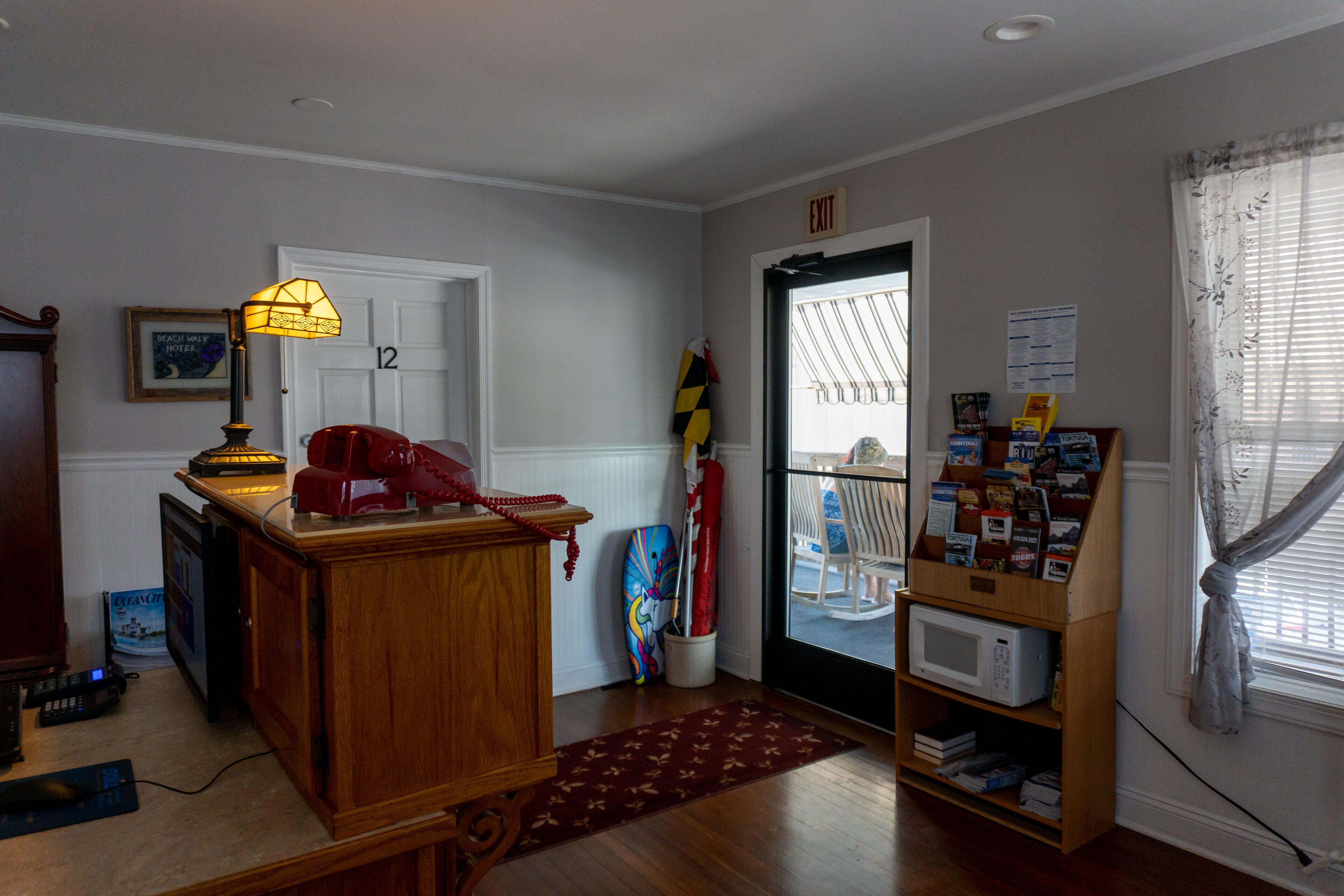 Una habitación con un escritorio de madera, una lámpara, una tabla de surf, una puerta con letrero de salida, una ventana con cortinas y un estante con revistas.