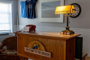 A reception desk with a wooden sign reading "WELCOME TO THE BEACH," a red telephone, a lamp, and a clock on the wall. - Beach Walk (Ocean City)