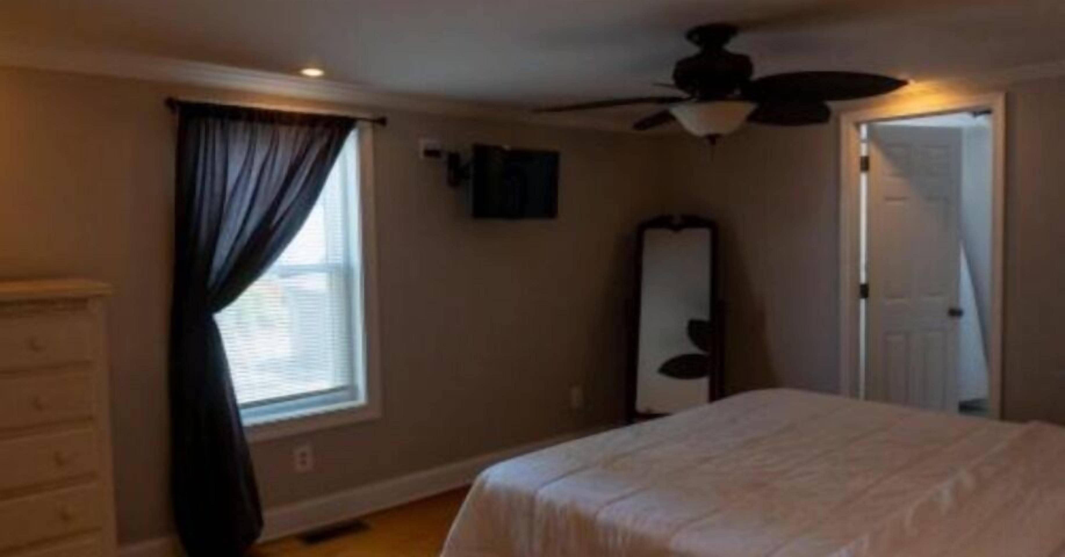 A bedroom with a bed, a dresser, a ceiling fan, a window with curtains, and a door.