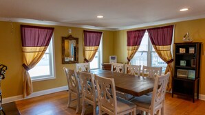 A dining room with a wooden table and six chairs, large windows with purple curtains, and a wooden bookshelf.