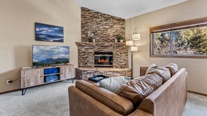 A living room with a stone fireplace, a leather sofa, a television, and a window with a view of trees. - Contemporary Country Club Condo with portable A/C's in the bedrooms. (Flagstaff)