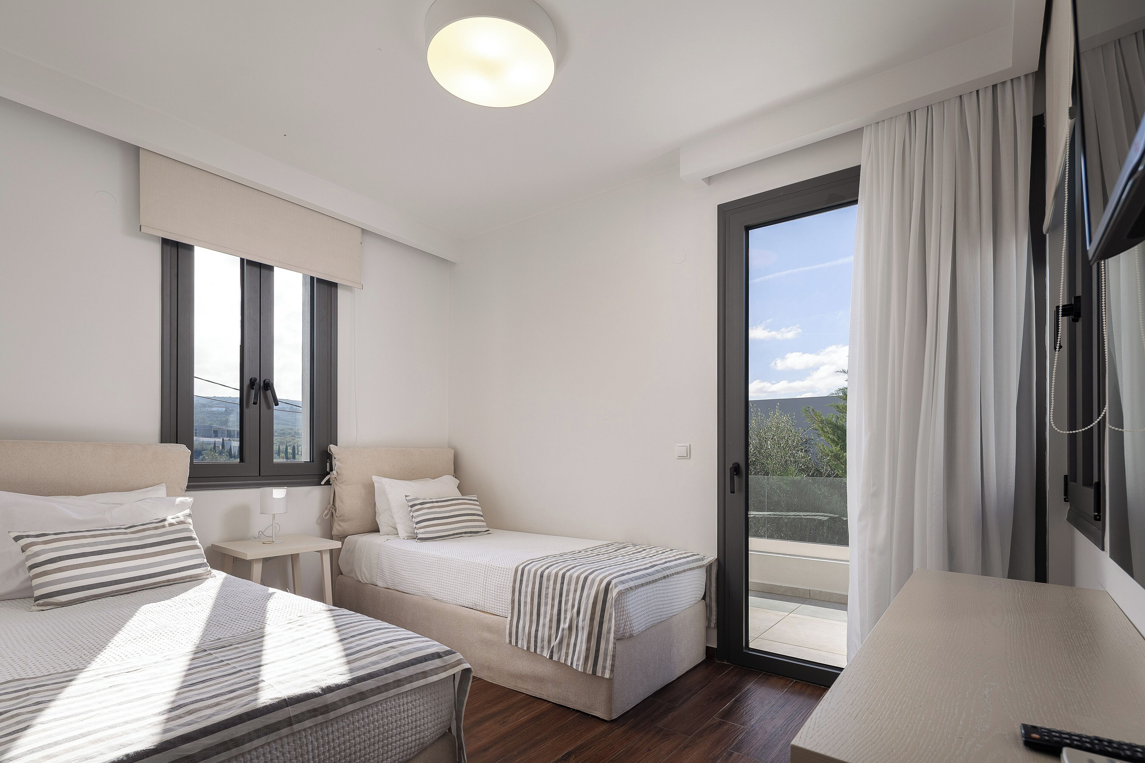 A modern hotel room with two beds, a small table, a TV, and a balcony with curtains.