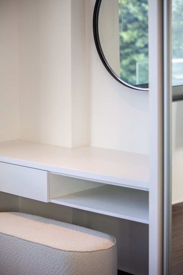 A white desk with a mirror, a small bench, and a window with a view of trees.